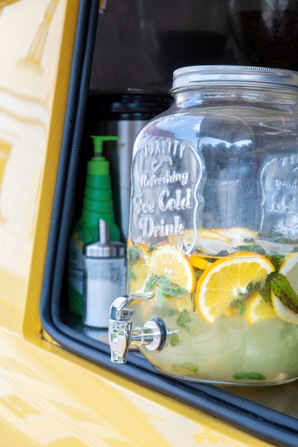 A glass jar with tap full of lemonade with lemon and mint in yellow food truck, close-up. Summer drink with ice in mobile retro coffee shop.