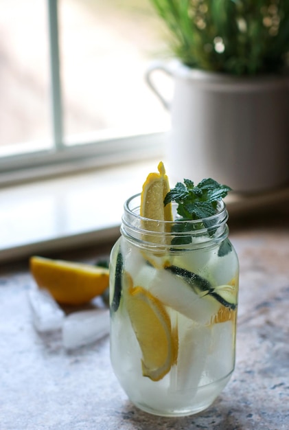 a glass jar with water, lemon, cucumber and mint on a marble table, blurred background