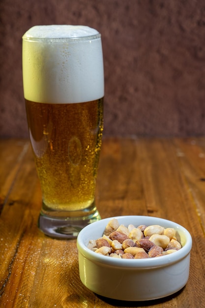 Glass of lager beer with peanuts on the table and in a white bowl on a wooden table.