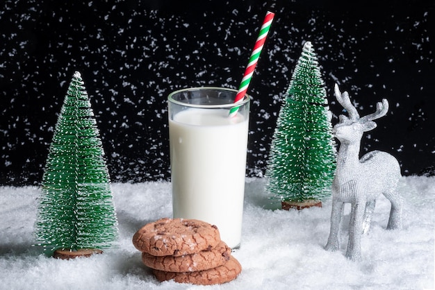 A glass of milk with a straw and cookies with chocolate chips on a snowy background among toy Christmas trees