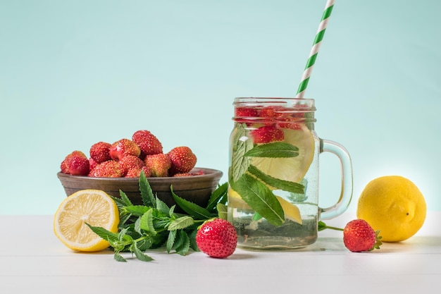 A glass mug of cold water with lemon mint and berries on a white wooden table