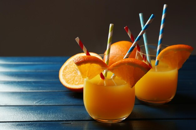 Glass of orange juice with straws and slices on color wooden table on dark background