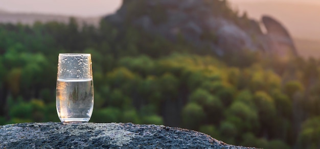 A glass of pure transparent mineral drinking water against the background of a sunset in the mountai...