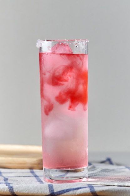 A glass of red cocktail with ice cubes and a blue spoon on a wooden board.