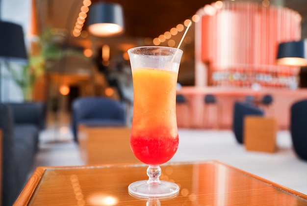 A glass of red and orange cocktail sits on a table in front of a bar.