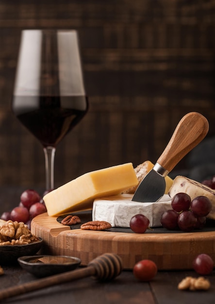 Glass of red wine with selection of various cheese on the board and grapes on wooden table background Blue Stilton Red Leicester and Brie Cheese and knife with honey