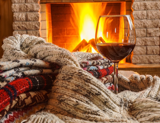 Glass of red wine and wool things near cozy fireplace. 