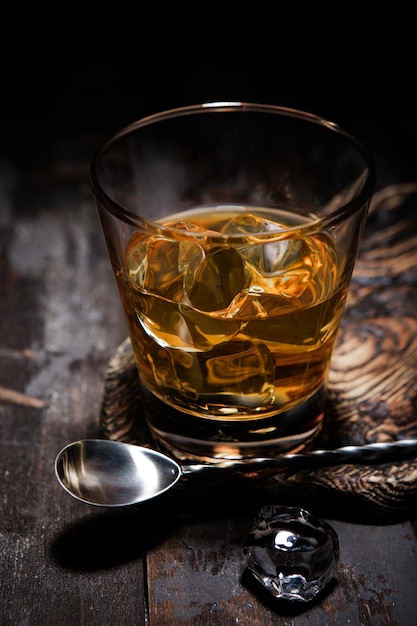 Glass of single malt whiskey with bar spoon and ice cubes on wooden background