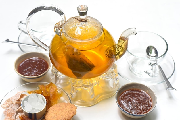 Glass tea pot with cups marmalade and crystalized fruits on white background