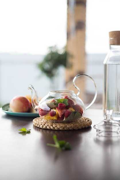 A glass teapot with fruits and berries mint leaves a bottle of water on the table outside Summer fruit tea