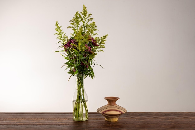 Glass vase with bouquet of beautiful flowers near wood vase over wood table on white wall background