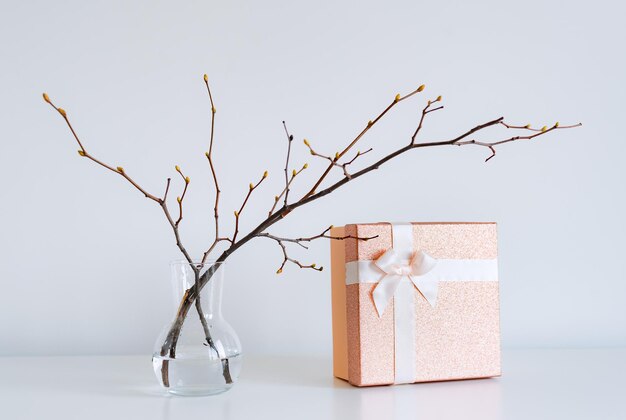 Glass vase with spring tree branches green buds and golden gift box on a white background Minimalism