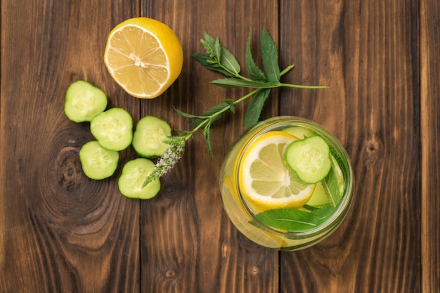 A glass of water with lemon and cucumber and cucumber slices on a wooden background