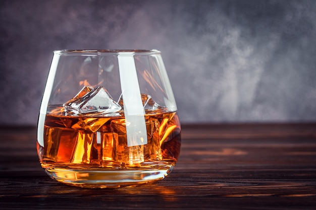 A glass of whiskey on the dark brown wooden table. Transparent yellow alcoholic beverage with ice. Brandy, bourbon. Strong alcohol drink. Copy space for text, template.