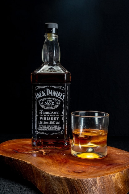 Glass of whiskey served with ice and bottle of Jack Daniels on rustic wood on black background.