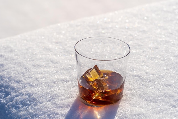 Glass of whiskey with ice on a bed of snow and white background close up