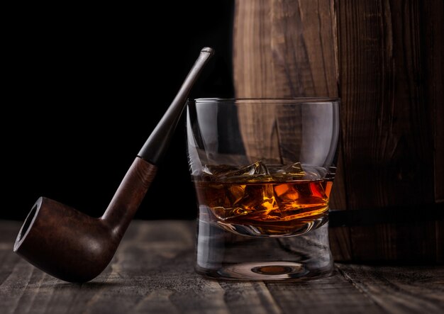 Glass of whiskey with ice cubes and vintage smoking pipe next to wooden barrel