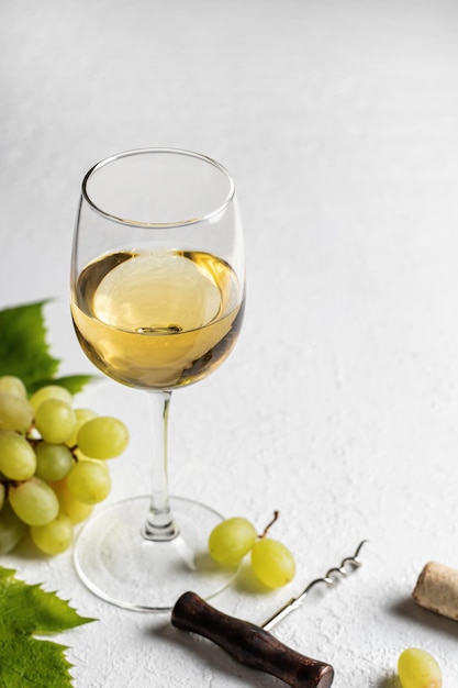 A glass of white wine on white textured background with corkscrew and grapes with text space