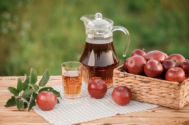 A glass with apple juice and basket with apples on wooden table with natural orchard background Vegetarian fruit composition