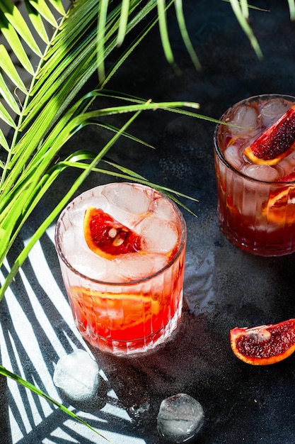 Glass with cold mineral water with blood oranges and ice cubes on concrete background Tropical palm leaf shadow Sunlight and shadow background