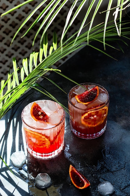 Glass with cold mineral water with blood oranges and ice cubes on concrete background Tropical palm leaf shadow Sunlight and shadow background