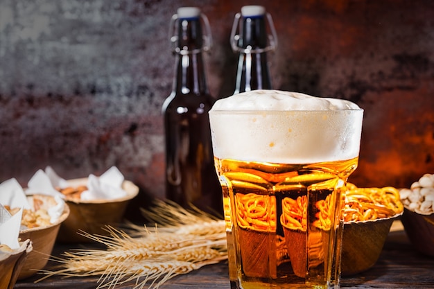 Glass with freshly poured beer and large head of foam near bottles, wheat and plates with snacks on dark wooden desk. Food and beverages concept