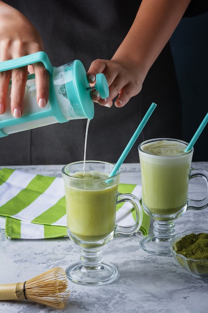 Glass with green latte. Matcha green tea and soy milk drink. Girl's hands add milk to a glass, cropped photo