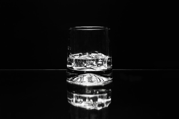 Glass with ice cubes in clear liquid on black background with reflection