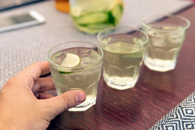 A glass with lemonade lemon wedges ice cubes Summer cold refreshing drink in the hand of a man on the table in a cafe 3 glasses of cool refreshing lemonade are on a wooden table in a summer cafe