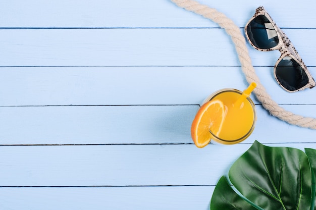 Glass with orange juice, monstera leaf and sunglasses on blue boards, copy space