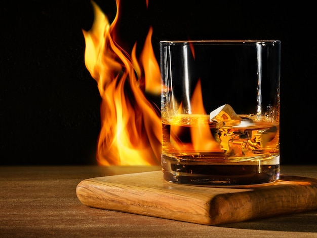 Glass with whiskey with ice on wooden table and black background with fire