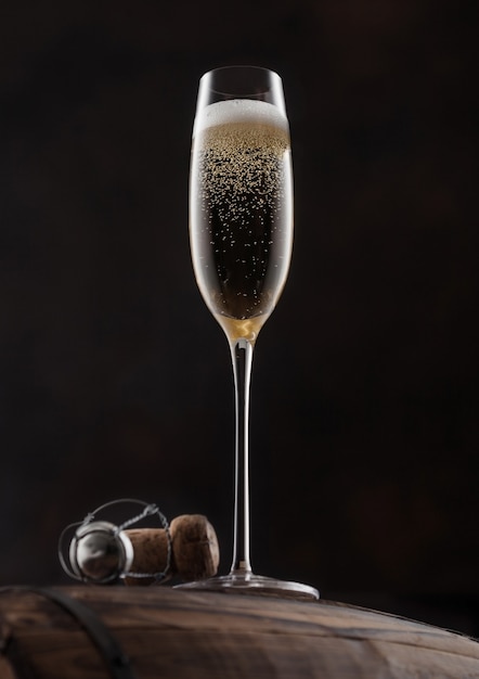 Glass of yellow champagne with cork and wire cage on top of wooden barrel on black background.