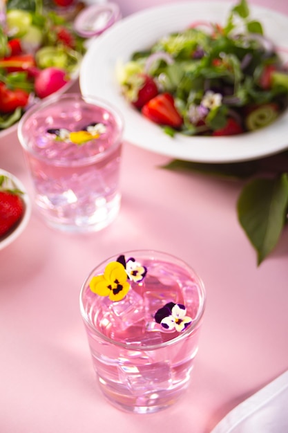 Glasses of pink violet purple cocktail decorated flowers viola pansy and fresh salad on the background