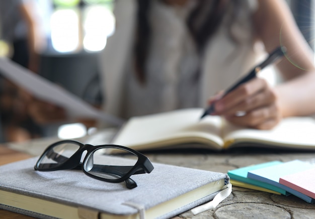 Glasses placed on a notebook, blurred women are checking graphs and writing notes.