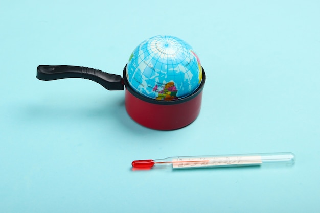 Global warming concept. Mini globe in a toy pan and thermometer on blue wall Minimalism. Climatic problems of our time.