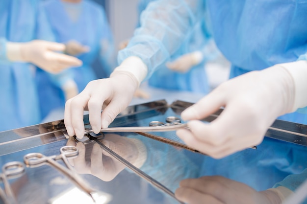 Gloved hands of surgeon or assistant in uniform taking one of sterile instruments from steel tray during operation