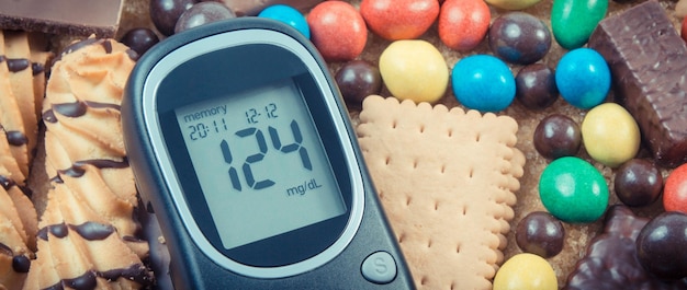 Glucose meter for checking sugar level and heap of sweets containing a lot of sugar Unhealthy and caloric eating