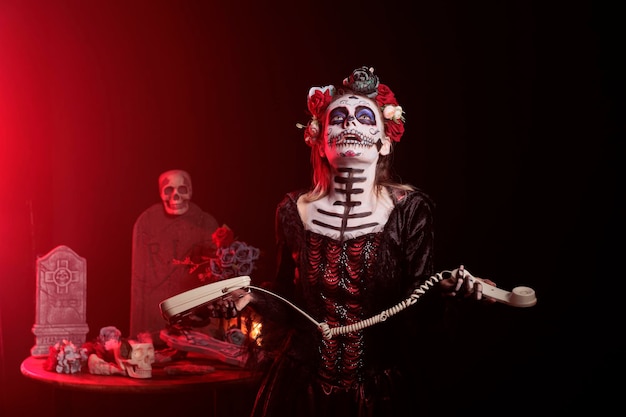 Goddess of death holding landline phone in studio, wearing skull make up and santa muerte costume. Woman using office telephone with cord and celebrating holy mexican ritual culture.