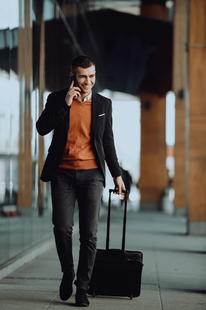 Going to the airport terminal. Young confident businessman traveler walking on city streets and pulling his suitcase drinking coffee and speaking on a smartphone. High quality photo