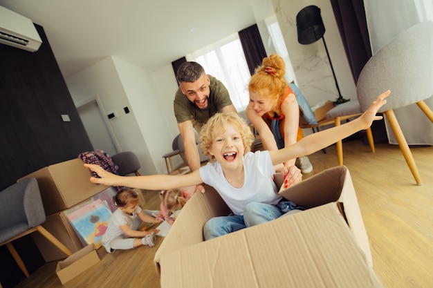 Going forward. Positive happy man pushing the carton box with his son in it while having fun