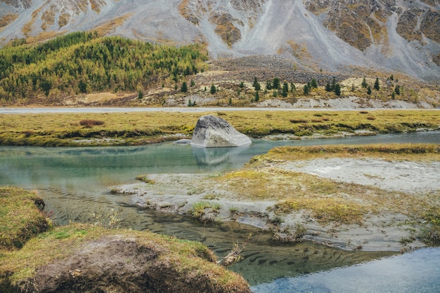 Golden autumn landscape with big stone in mountain creek with view to forest hillside. Beautiful big boulder in turquoise clear water stream in autumn. Green ripple sandy bottom in transparent water.