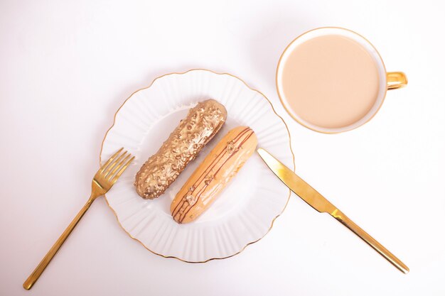 Golden colored eclairs on plate with golden knife and fork and a cup of coffee on white background