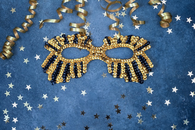 Golden glittering mask and serpentine with golden stars on blue background top view copy space