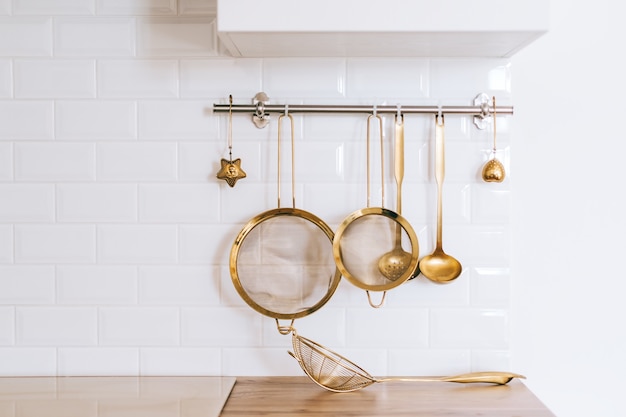 Golden kitchen utensils for cooking on a white wall with copy space.