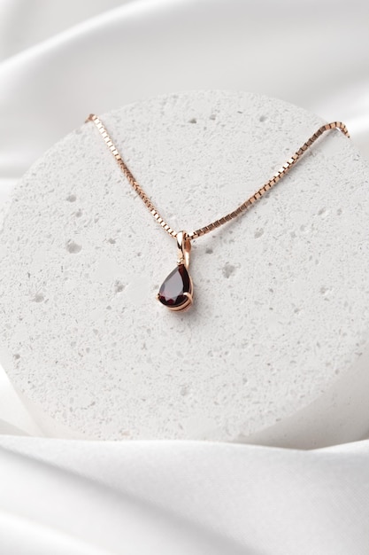 Golden necklace with garnet on concrete podium on white silk background rose gold Beautiful accessories for women Elegant jewelery gift or present for wedding or saint valentine's day