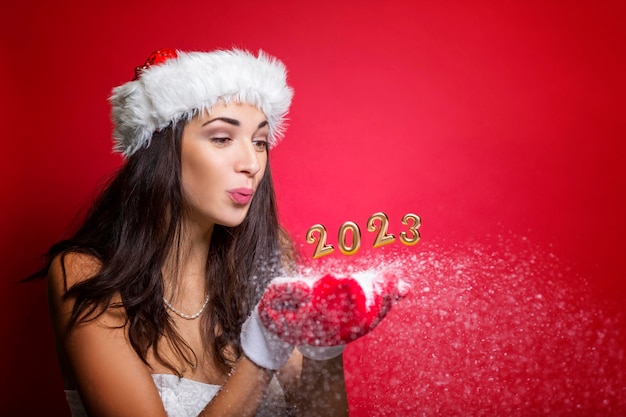 Golden numbers 2023 with snow on the palms of a young woman in red mittens and a santa hat Beautiful festive brunette in a white dress Magic for Christmas and New Year Red background