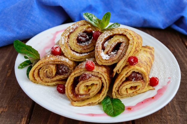 Golden pancakes in the form of roll with strawberry jam and powdered sugar on a white plate on a wooden table. Close-up. Breakfast