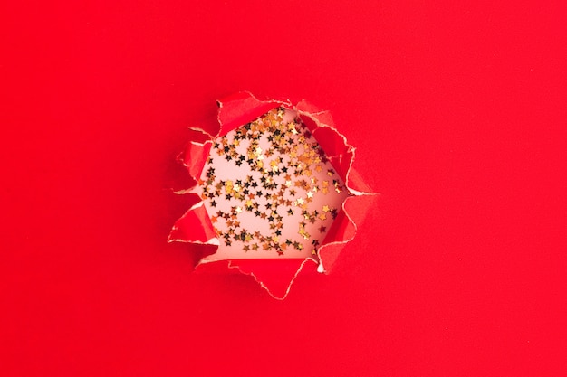 Golden stars confetti through a hole in red background. Minimal festive concept.