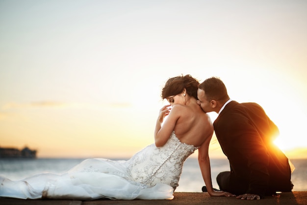 Golden sun shines behind groom kissing bride's shoulder while they rest on the shore