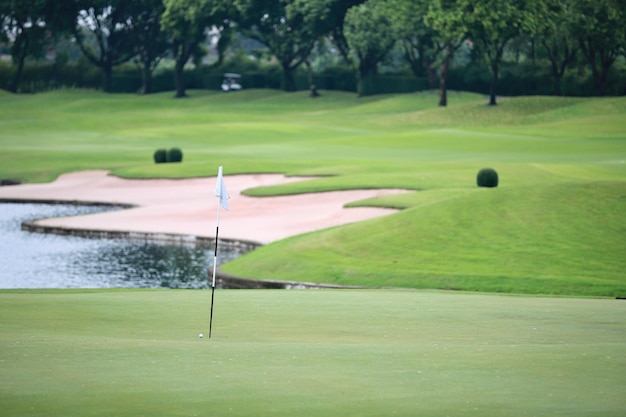 A golf ball that almost hits the hole with the flag On the green with many signs of repair In the background are sand bunkers ponds and lush lawns There is a blurred golf cart in the distance